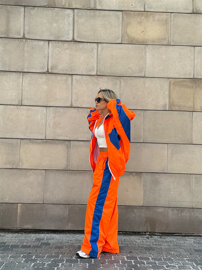 Model in a bright orange windbreaker set with royal blue panels and a wide blue stripe down the trousers, standing against a stone wall. 15