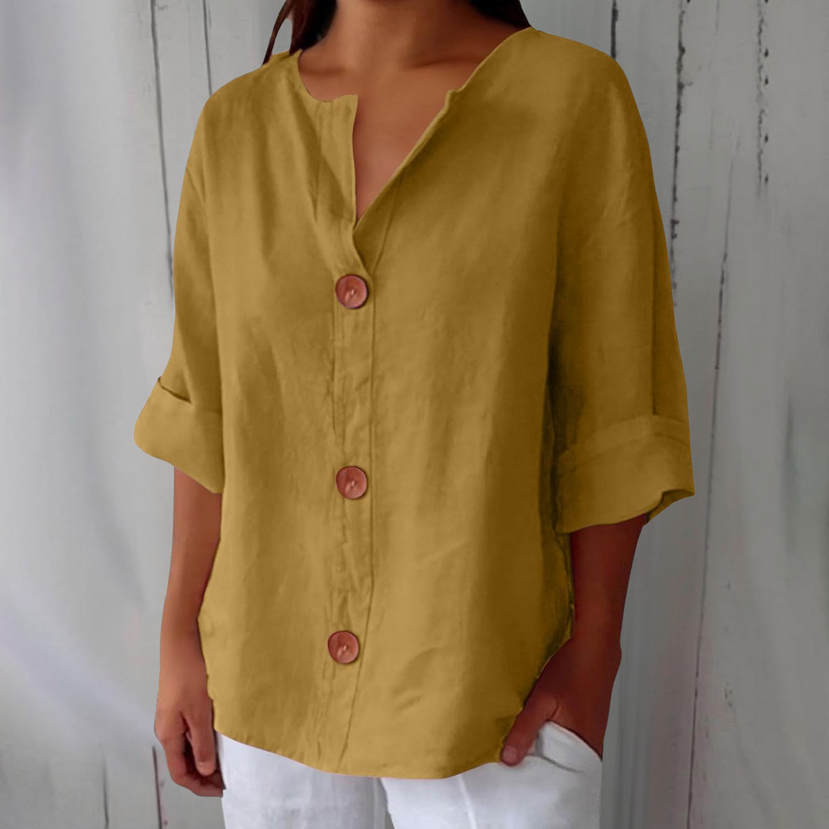 Close three-quarter view of the loose blouse in mustard yellow, with notched V-neck, three brown buttons and cuffed sleeves, hand in trouser pocket. 6