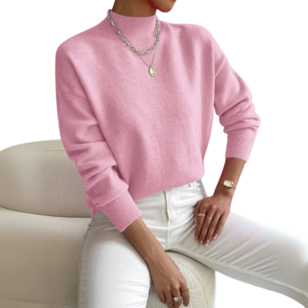 Model seated on a cream chair wearing the jumper in pink with white jeans, layered silver necklaces and a gold watch. 1