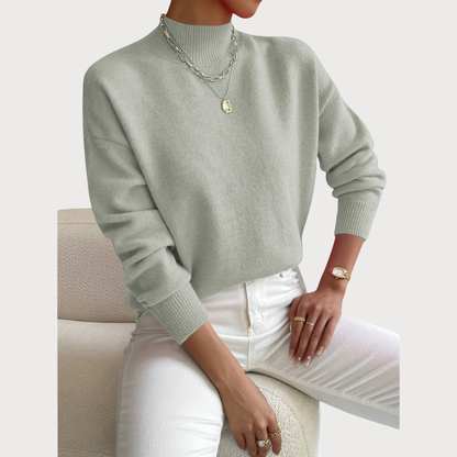 Jumper in soft sage green worn by a seated model with white jeans, with a ribbed high neck and cuffs. 2