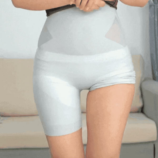 Model in light grey high-waisted shaping shorts on a sofa, tugging the top of the waistband, front view. 3