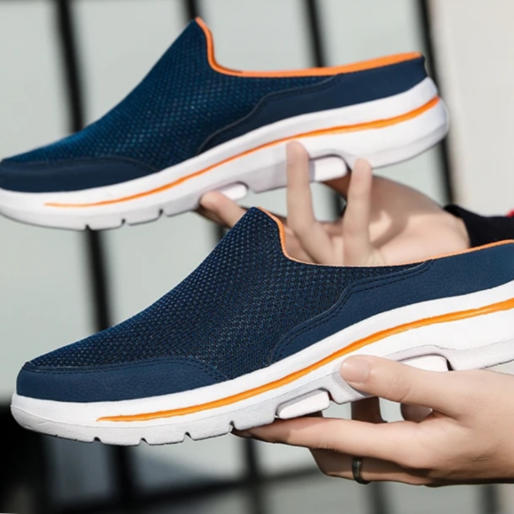 Two hands holding a pair of navy mesh slip-ons with orange trim and white soles, showing the side profile. 12
