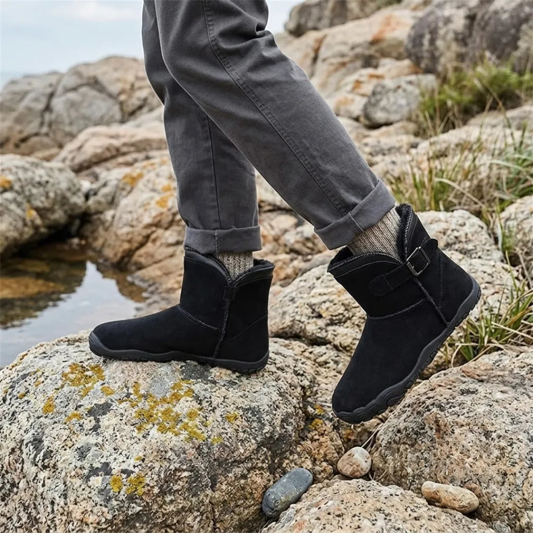 Black boots worn on lichen-covered rocks beside water, with rolled grey trousers and wool socks 11