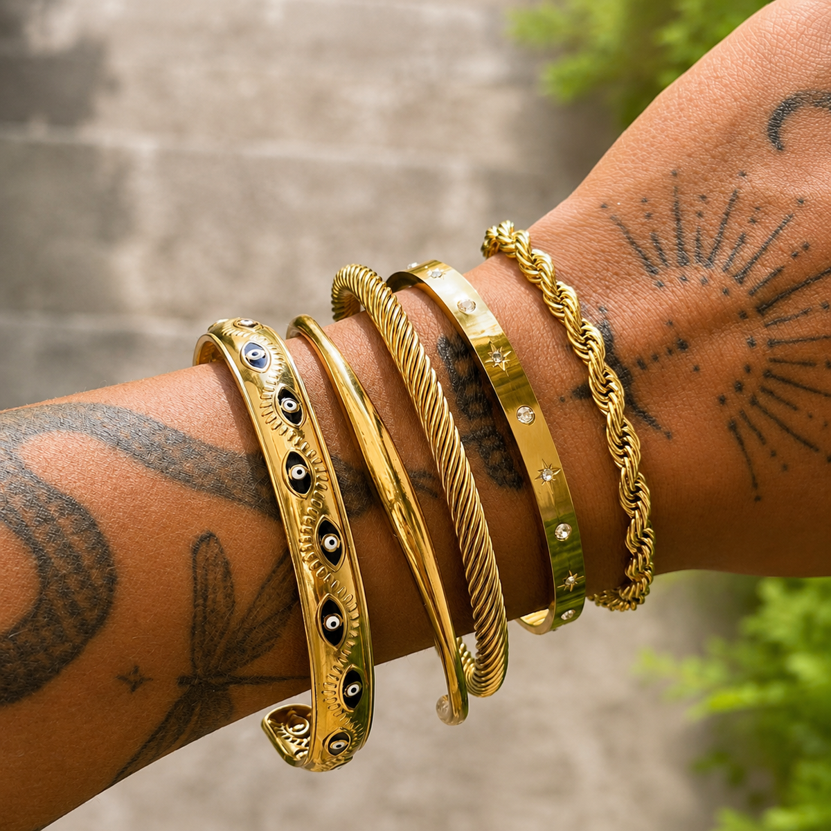 Slim gold cuff worn on a tattooed wrist, stacked with four other gold bangles and bracelets, including twisted, eye-motif and crystal-studded designs. 3