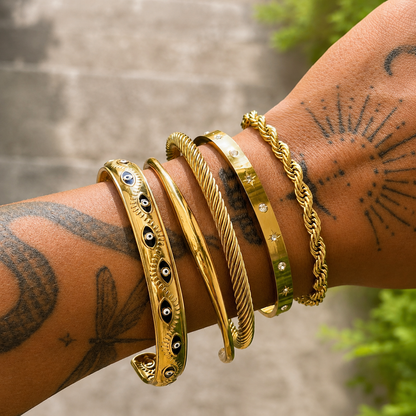 Slim gold cuff worn on a tattooed wrist, stacked with four other gold bangles and bracelets, including twisted, eye-motif and crystal-studded designs. 3