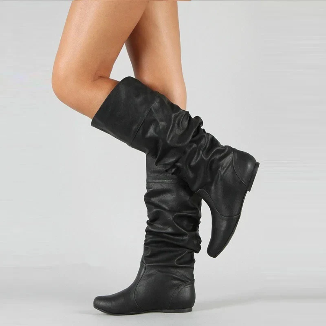 Model's bent leg in black slouchy flat boots with a folded cuff and ruched shaft, shown from the side. 3