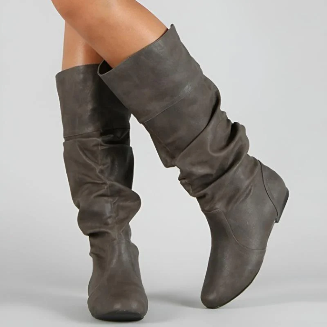 Close view of grey-brown slouchy flat boots worn by a model, showing the ruched shaft, folded cuff and rounded toe. 4