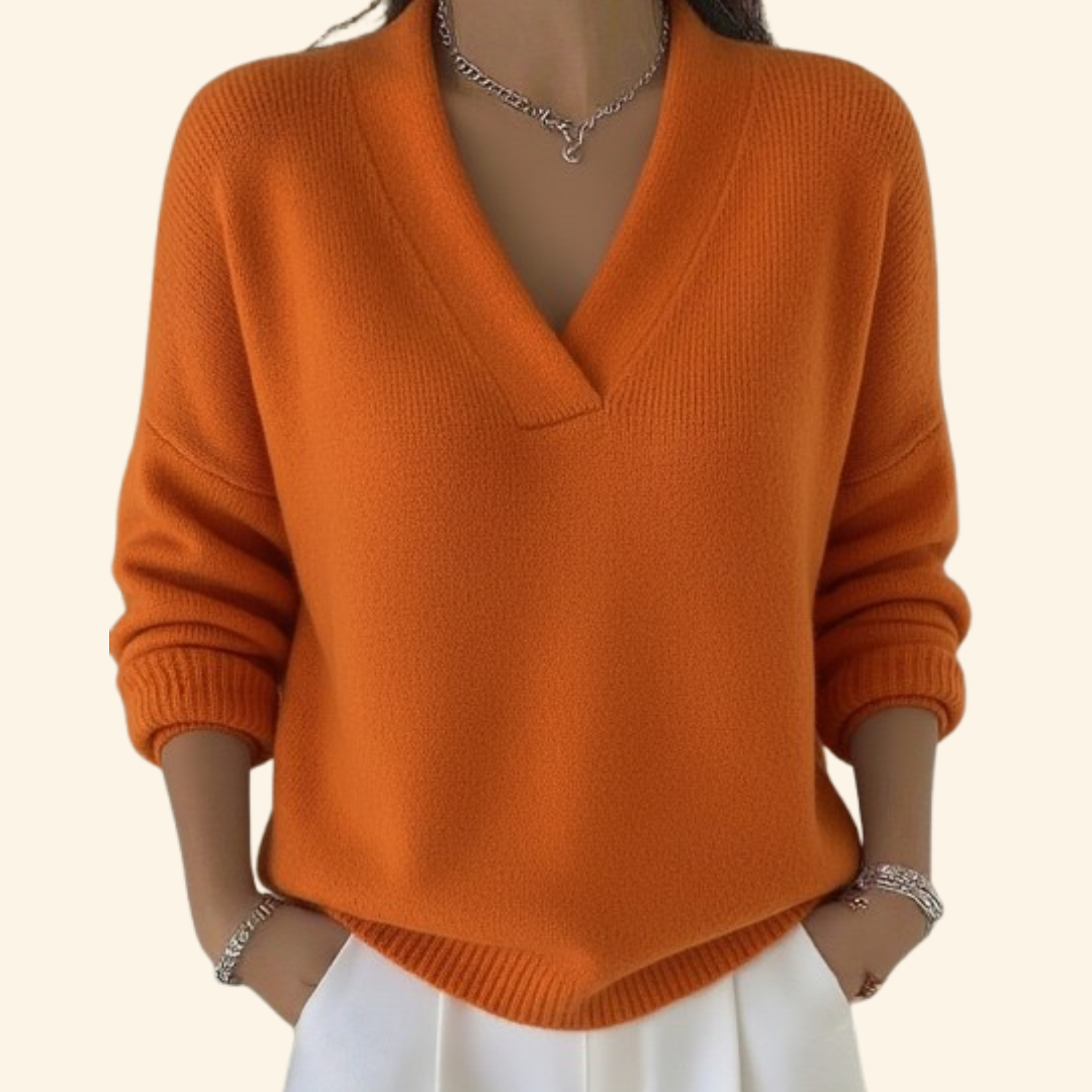Model wearing the loose V-neck knit in bright orange with white trousers, silver necklace and bracelets, hands in pockets. 5