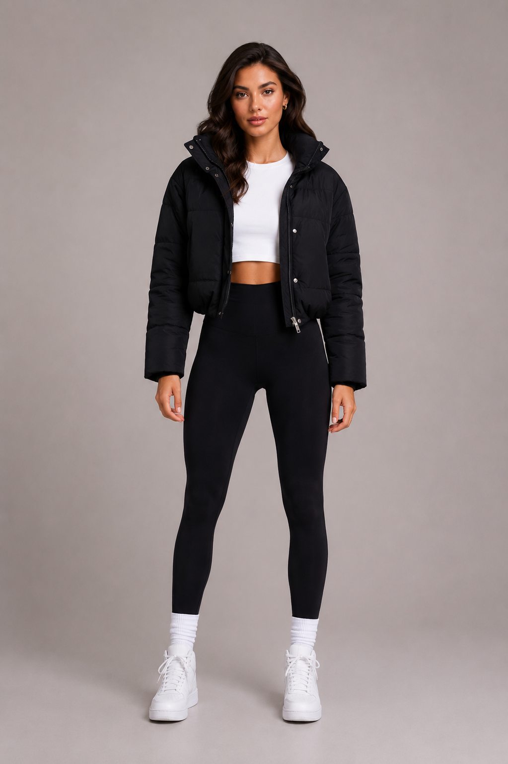 Full-length front view of a model in the black cropped puffer jacket with the hood down, styled with leggings and white trainers. 2