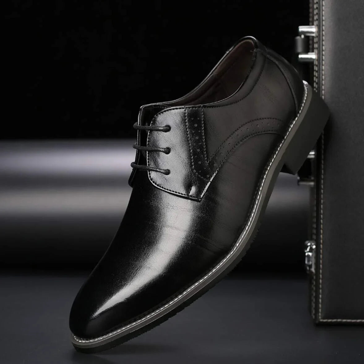 Single black polished lace-up shoe with brogue perforations and contrast stitching, shown side-on against a dark briefcase. 3
