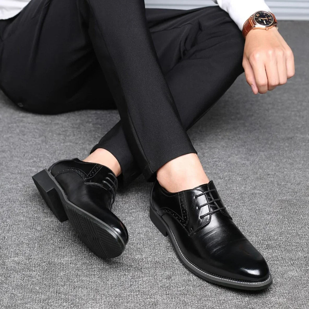 Man seated on grey carpet in black trousers wearing black lace-up shoes, one sole turned up to show its tread. 4
