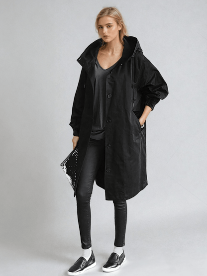 Model in the black hooded coat with skinny trousers and slip-on trainers, holding a clutch 2