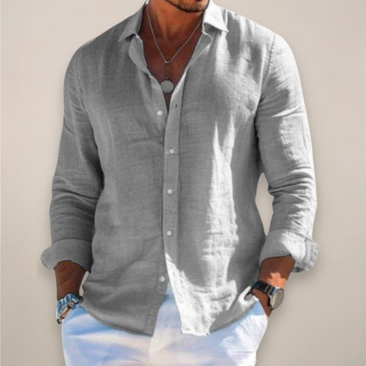 Model wearing the grey version of the shirt, buttoned with sleeves rolled up, hands in pockets of white trousers. 1