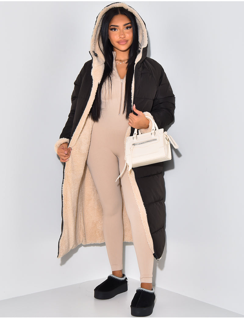 Model wears the black quilted side of the coat open, showing the cream teddy-fleece lining and hood, with a beige jumpsuit and white handbag. 1