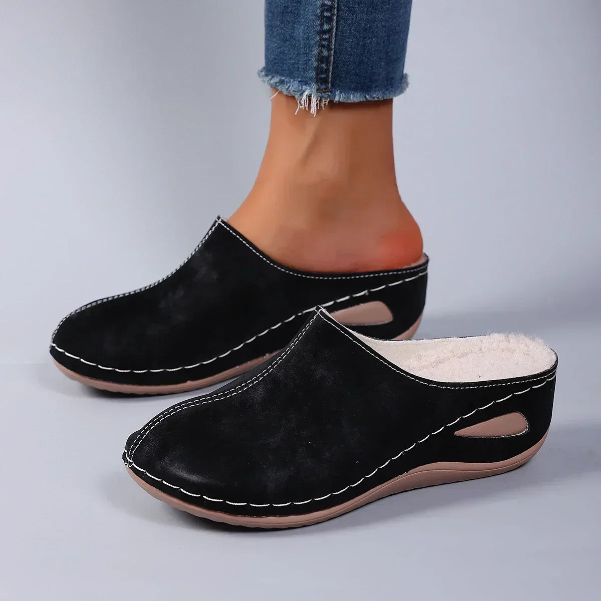 Black backless wedge slippers worn with frayed jeans, side view showing white stitching, cut-out detail and fleece lining at the opening. 2