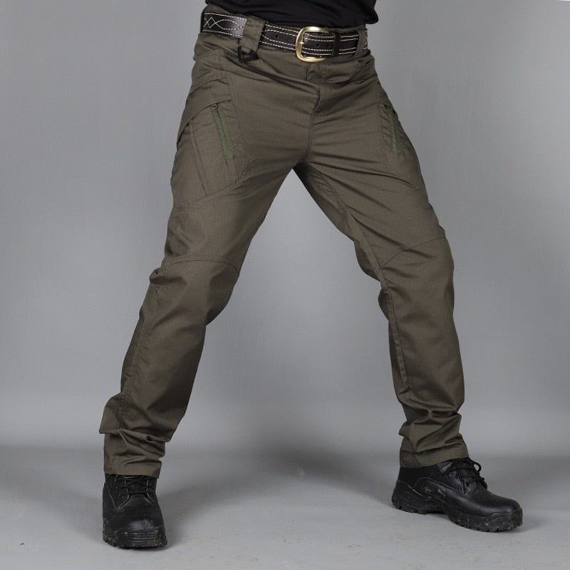 Front view of olive green trousers worn with a leather belt and black boots, showing zipped thigh pockets against a grey backdrop. 4