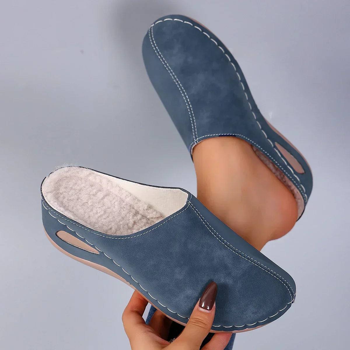 Blue backless slippers held up to show the soft cream fleece lining and white contrast stitching, with a bare foot inside the other. 1