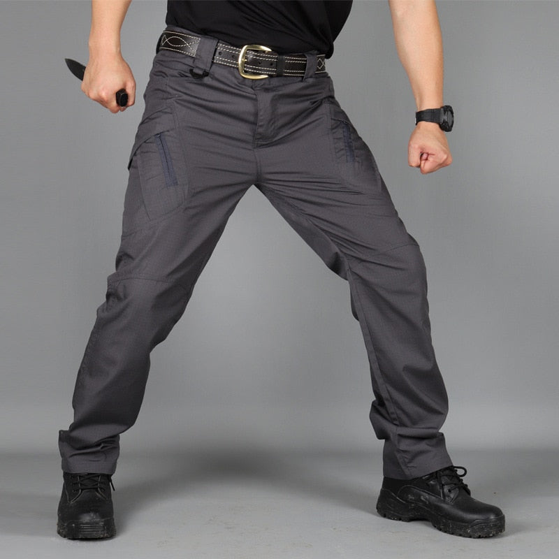 Front view of dark grey trousers worn with a leather belt and black boots, the model in a wide stance holding a knife. 6