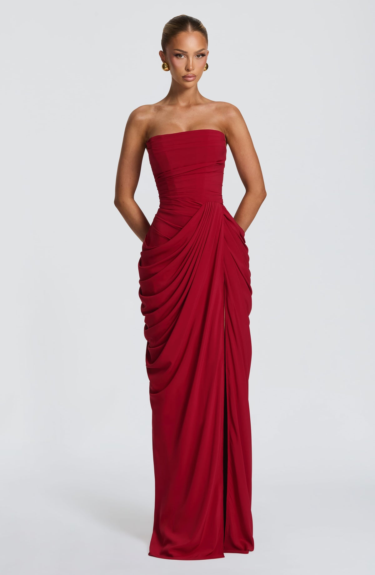 Model facing forward in the red strapless maxi dress with draped wrap skirt, hands in pockets and gold earrings. 27