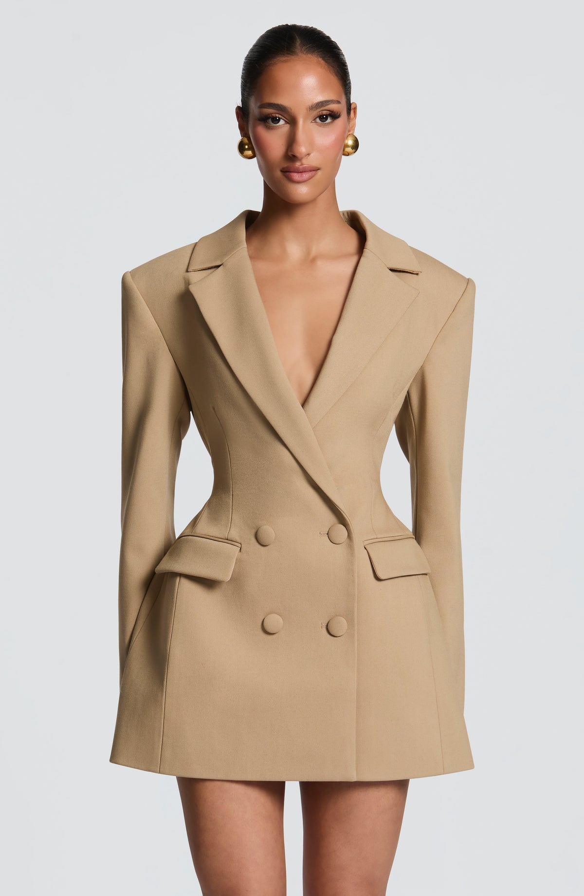 Front view of the camel-beige blazer dress with a deep plunge neckline, fitted waist, flap pockets and gold ball earrings. 18