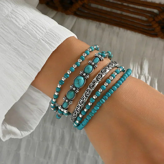 Five stacked turquoise bracelets with silver-tone beads and an ornate scrolled bangle, worn on a wrist with a white sleeve, seen from the side. 1