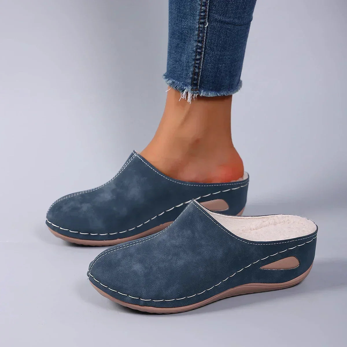 Blue backless wedge slippers worn with frayed jeans, side view showing the fleece lining, white stitching and teardrop cut-out in the sole. 4