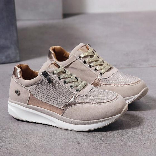 Pair of beige trainers with sparkly gold laces, a side zip and studded panels, shown on a grey floor. 2