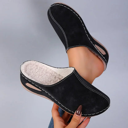 Black backless slippers held up by a hand with brown nails, showing the cream fleece lining and white stitching, one worn on a foot. 5