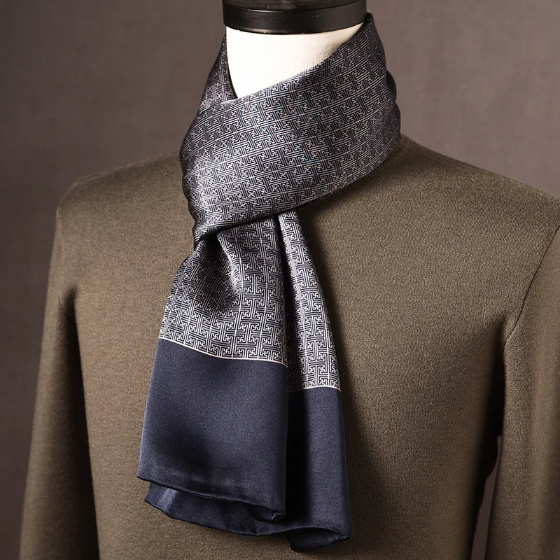 Grey and navy silk scarf with a fine geometric maze-like pattern and plain navy ends, draped on a mannequin over an olive jumper. 10