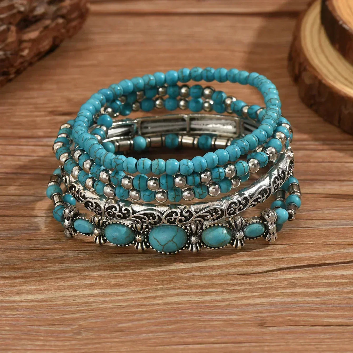 The five bracelets stacked on a wooden surface, showing the cracked turquoise beads, silver-tone beads and engraved scroll bangle up close. 3