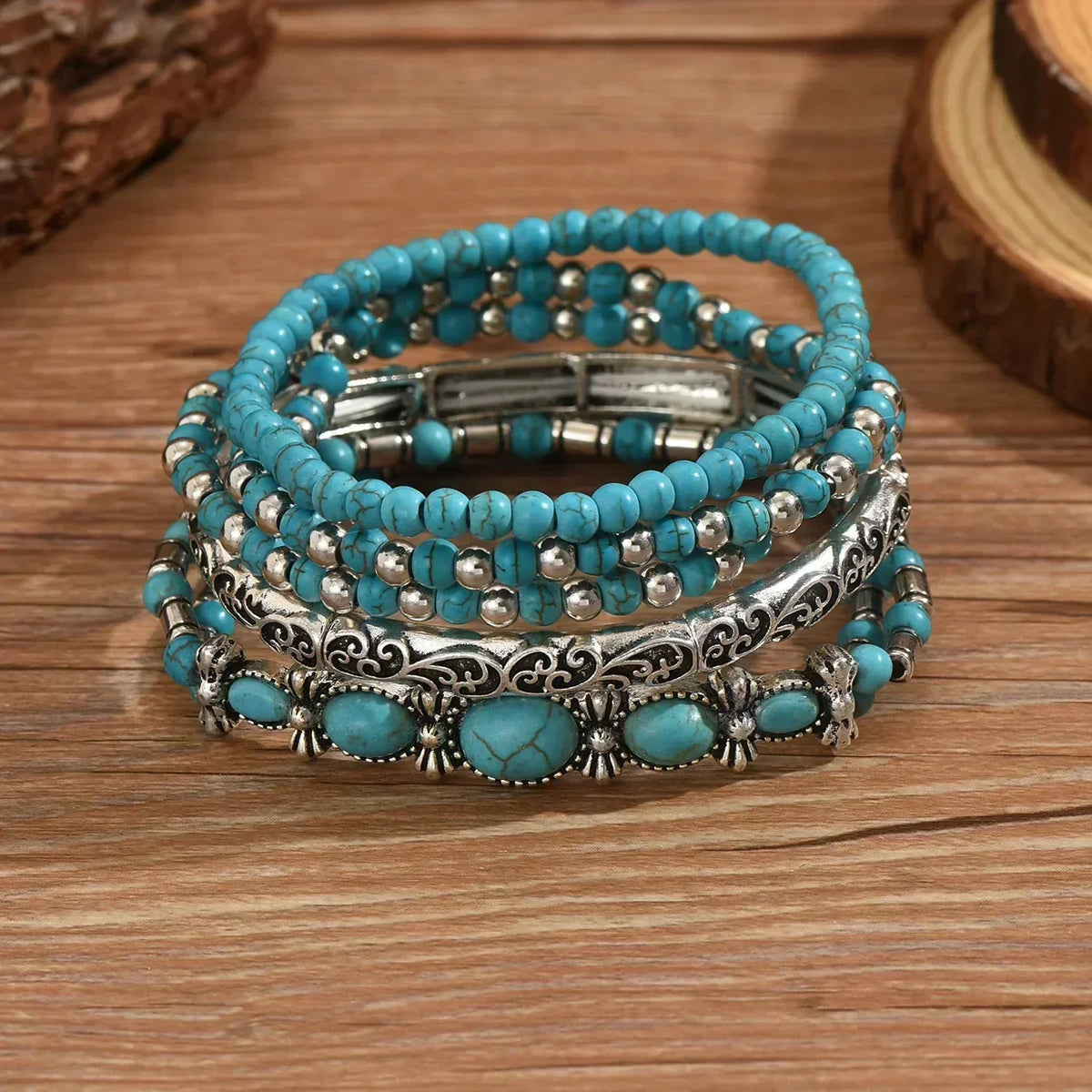 Stacked bracelets resting on a wooden surface, showing the oval turquoise-style stones, scrollwork silver-tone bangle and stretch beaded strands. 3