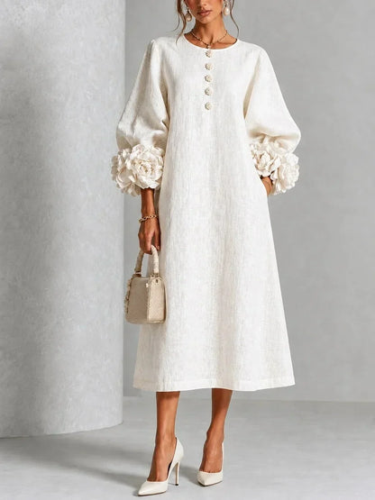 Model wears the cream long-sleeved midi dress with oversized fabric flowers at the cuffs and flower buttons down the front, holding a woven handbag. 1