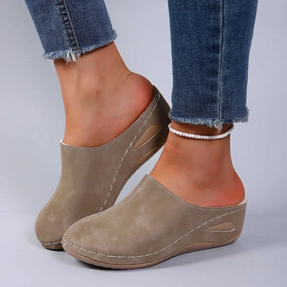 Taupe backless slippers worn with frayed jeans and a beaded anklet, showing the stitched upper, side cut-out and curved wedge sole. 3