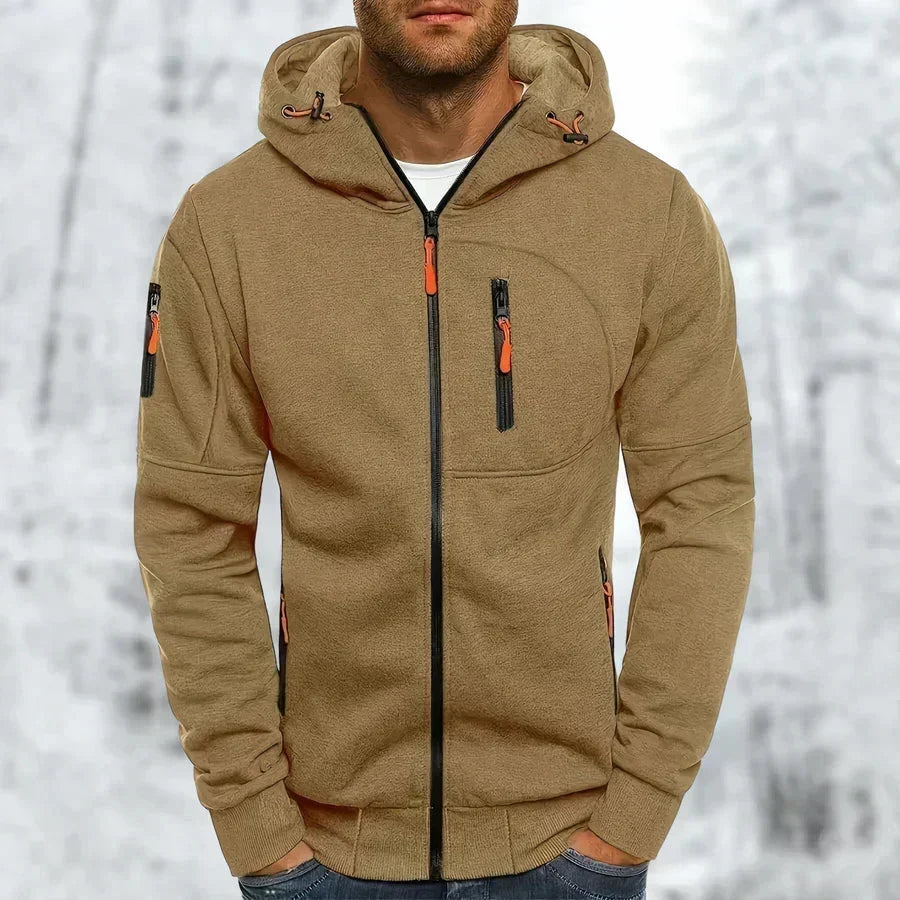 Model wearing the hoodie in tan khaki with orange zip pulls, black front zip and ribbed hem, in a snowy setting. 10