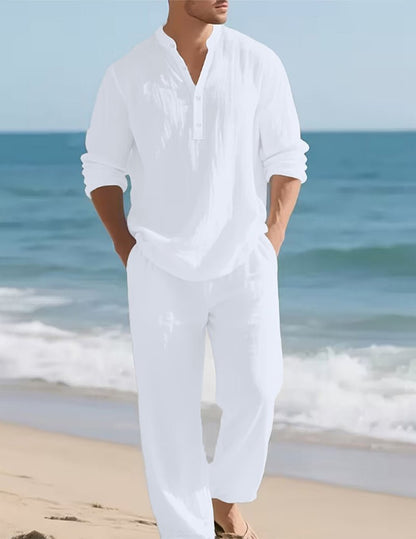 Model in the white set walking along a sandy beach with hands in pockets, waves and blue sea behind. 2