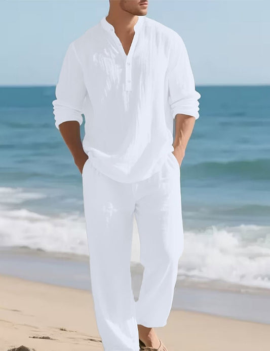 Model in the white set walking along a sandy beach with hands in pockets, waves and blue sea behind. 2