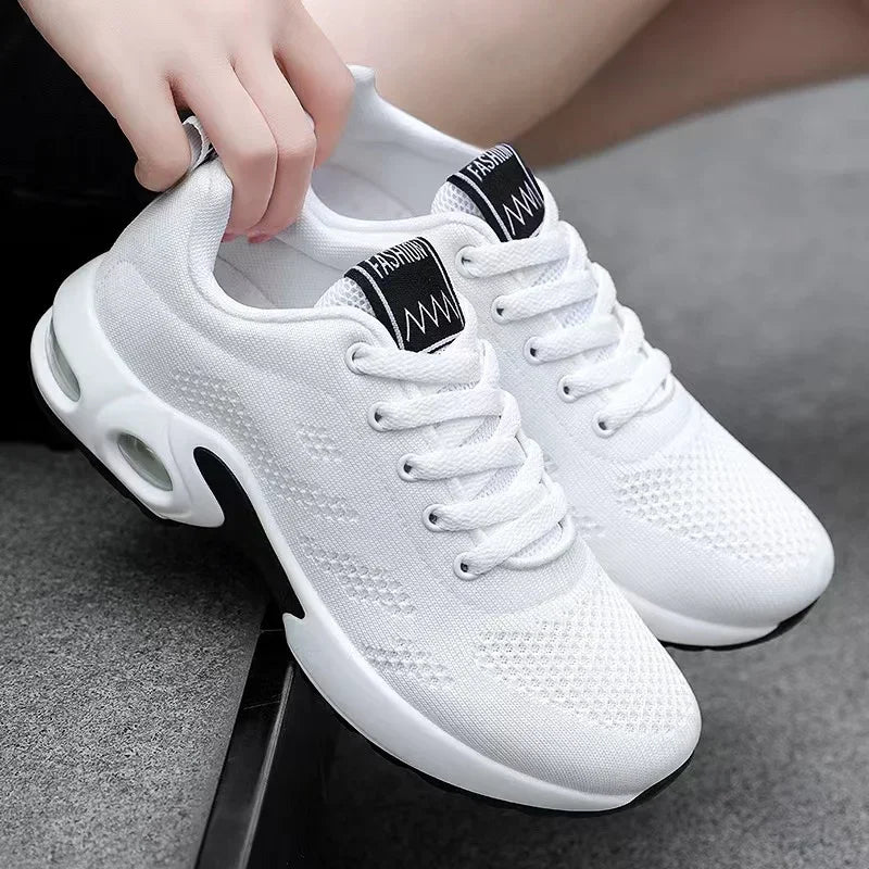 Pair of white knit lace-up trainers with white air-cushion soles, one held by a hand at the heel, set on grey steps. 2