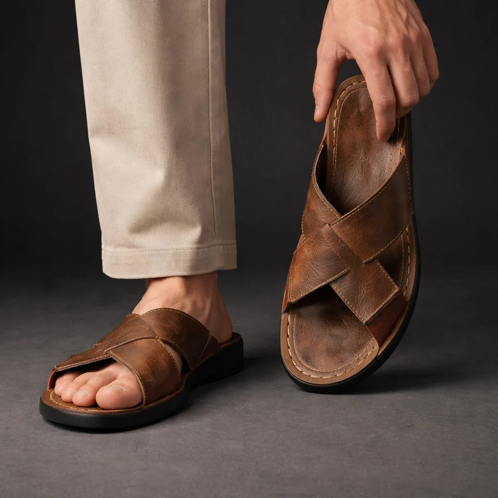 Man wearing a brown crossed-strap sandal with beige trousers, while holding the other sandal up to show its footbed and straps. 3