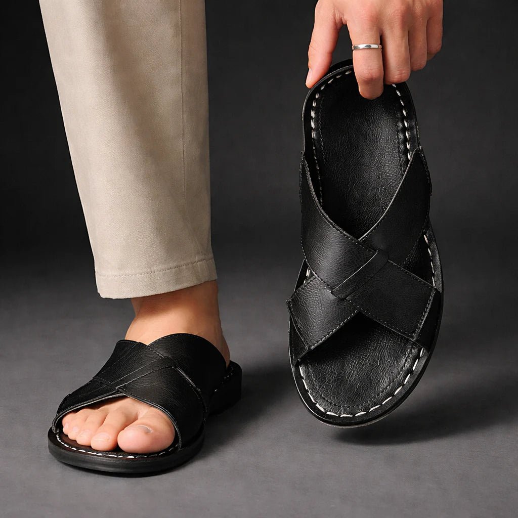 Man wearing a black crossed-strap sandal with beige trousers, while holding the other sandal up to show its stitched footbed. 7