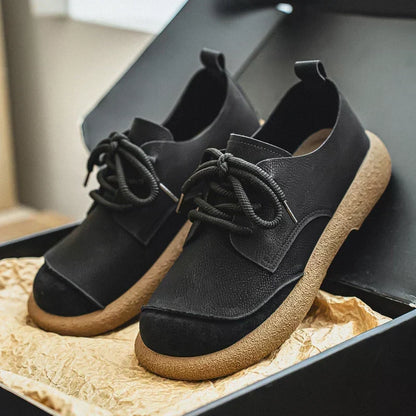 Black lace-up shoes with tan crepe-style soles and suede toe caps, displayed in an open box on crinkled paper. 12