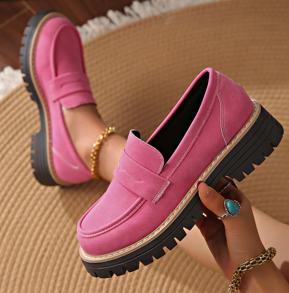 Bright pink suede-look loafer held in a hand in the foreground, with a second pair worn behind, on a woven rug. 18