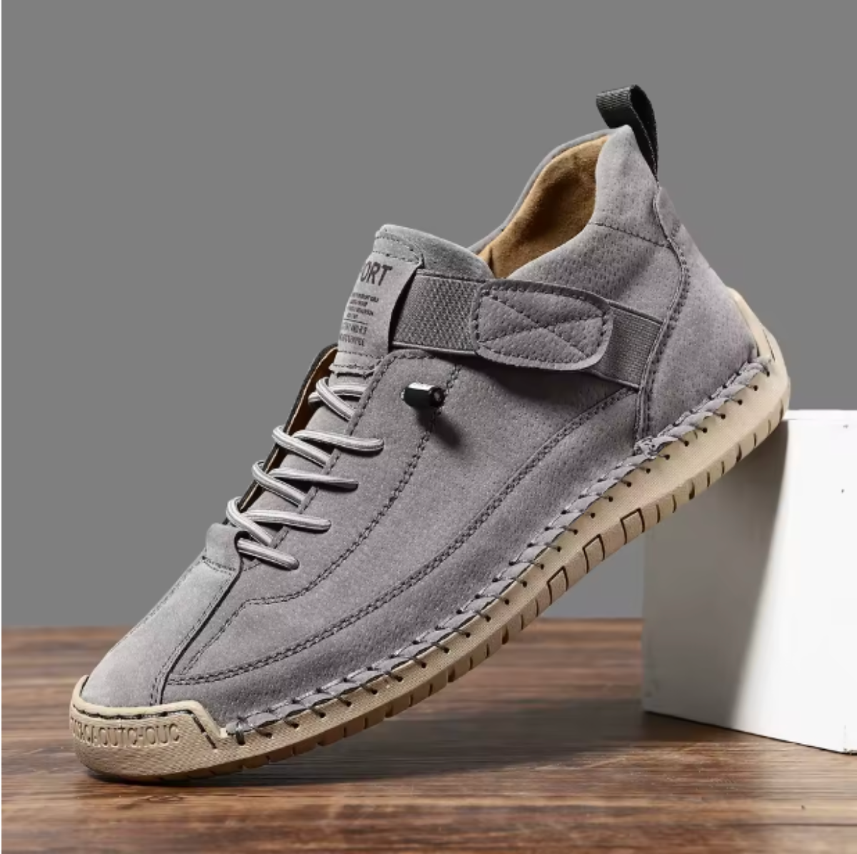 Single grey trainer with perforated finish, elastic strap and laces on a tan sole, angled against a white block. 10