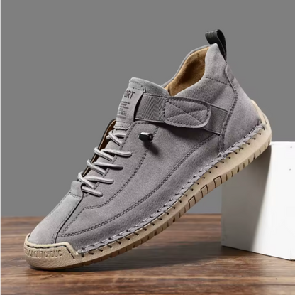 Single grey trainer with perforated finish, elastic strap and laces on a tan sole, angled against a white block. 10