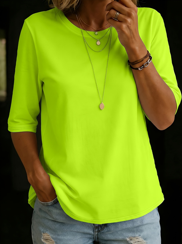 Woman wearing the round-neck half-sleeve t-shirt in bright neon lime green, with layered necklaces and ripped jeans, against a dark background. 5