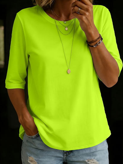 Woman wearing the round-neck half-sleeve t-shirt in bright neon lime green, with layered necklaces and ripped jeans, against a dark background. 5