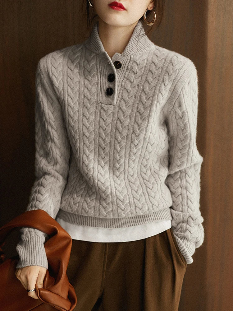 Model in the light grey cable-knit sweater with three dark buttons, white layer at the hem and brown trousers, holding a leather bag. 14