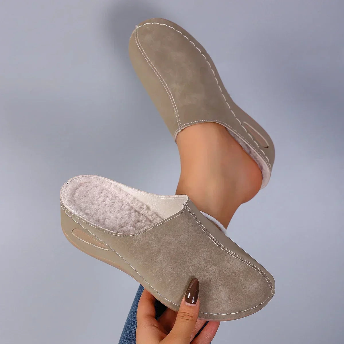 Taupe backless slippers held up to show the pale fleece lining and stitched upper, with the other slipper worn on a bare foot. 6