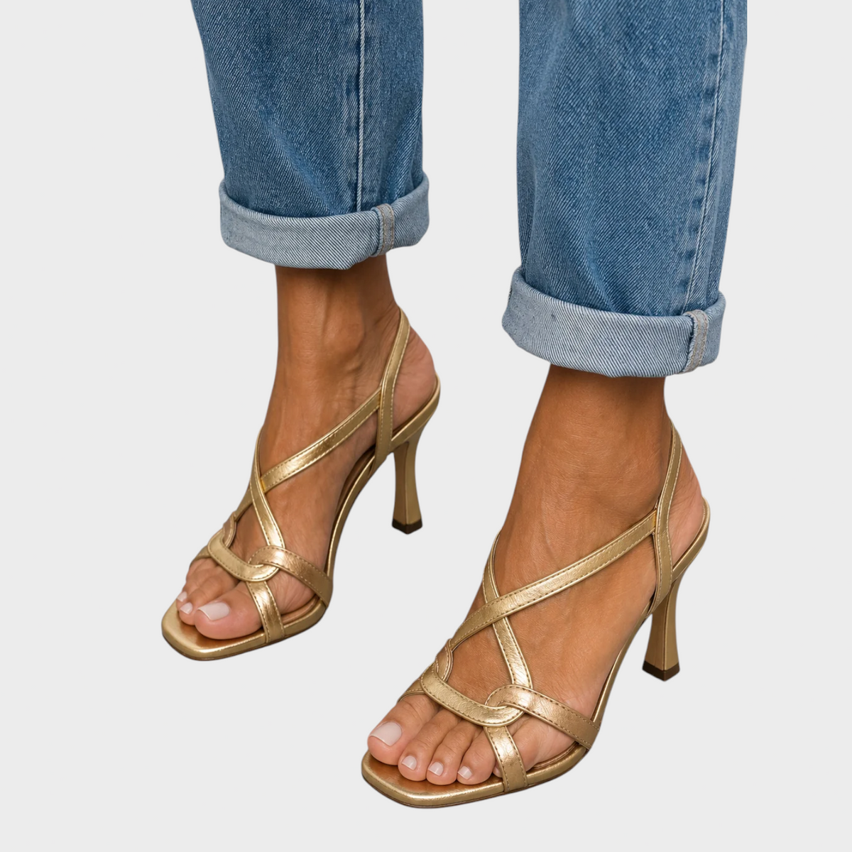 Close-up from the front of gold crossover-strap sandals with square open toes and flared heels, worn with turned-up blue jeans. 1