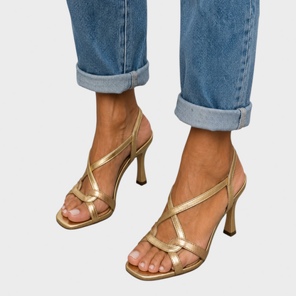 Close-up from the front of gold crossover-strap sandals with square open toes and flared heels, worn with turned-up blue jeans. 1