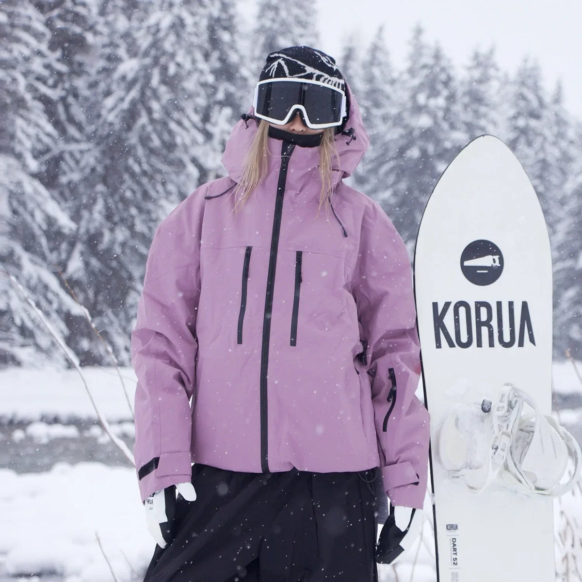 Model in a dusky pink jacket with black zips and cuff straps, standing beside a white snowboard in snowfall. 3